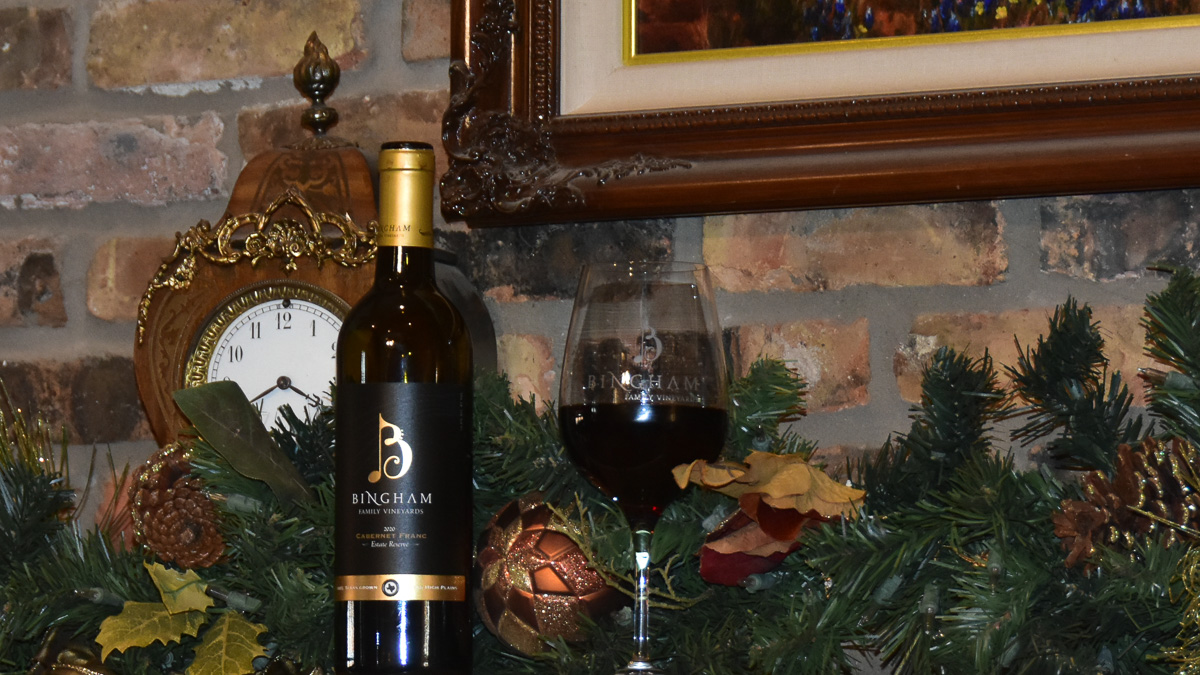 bottle of Bingham Family Vineyards 2020 Reserve Cabernet Franc with glass of wine on a fireplace mantel with Holiday decorations and a clock in the back ground