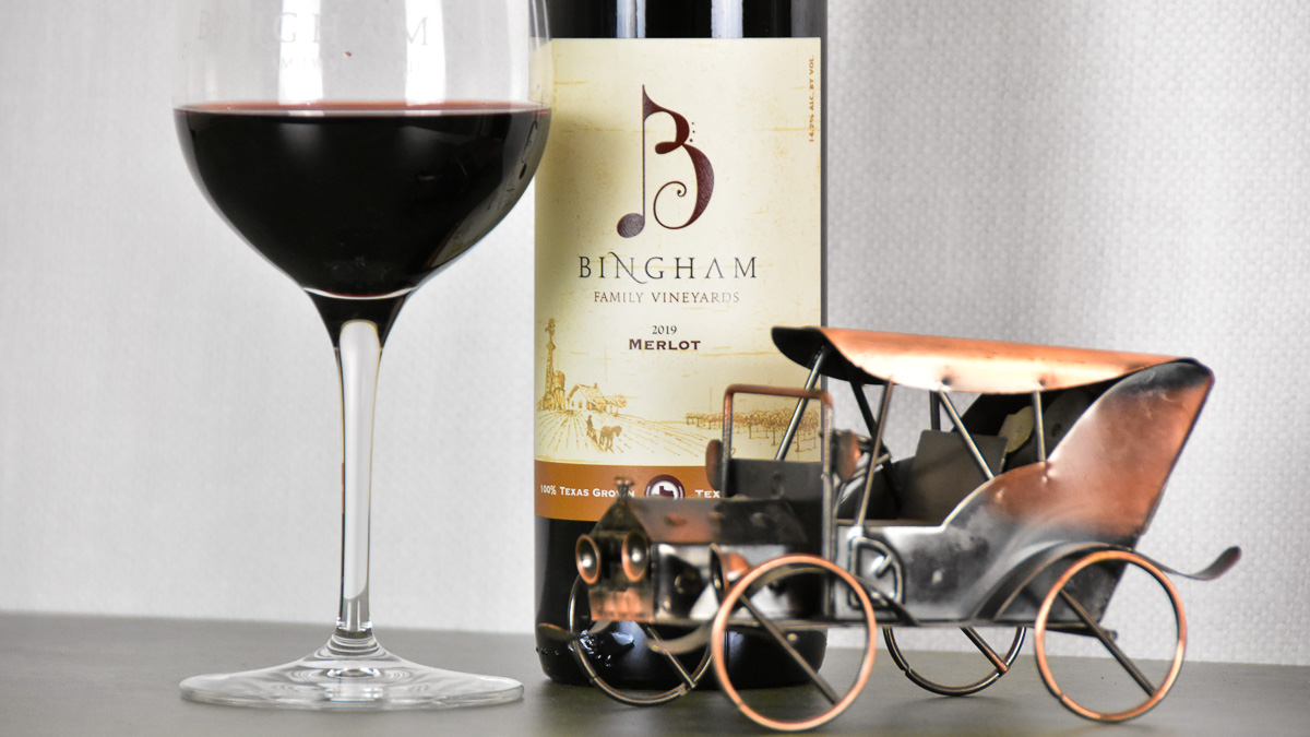 Bottle of Bingham Family Vineyards 2019 Merlot with a glass of wine and a miniature car model made of copper.