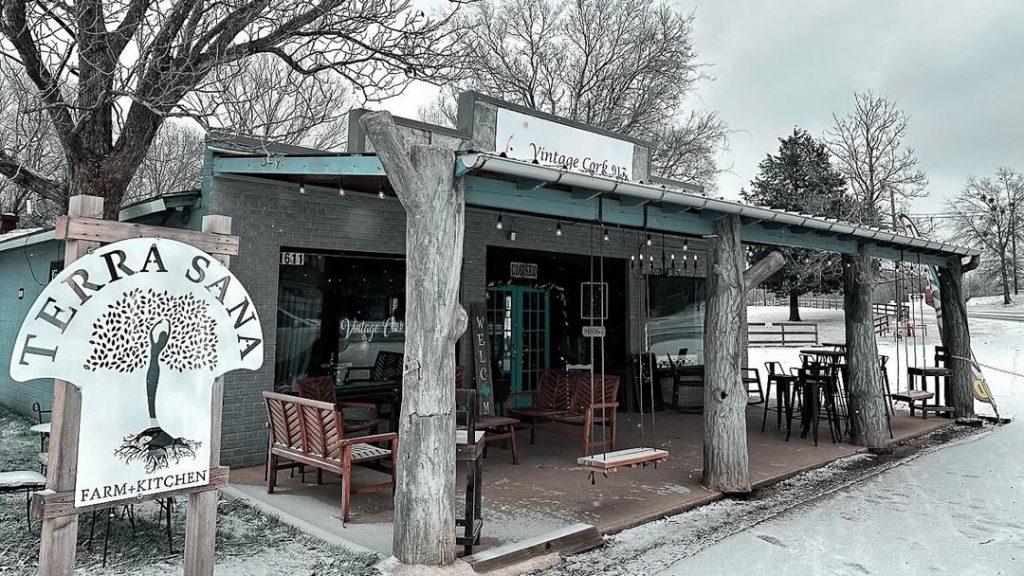 Outdoor cafe under a wooden covered porch with rustic chairs and a Terra Santa Farm Kitchen sign; snow on the ground. Vintage Cork, Ben Wheeler, Texas