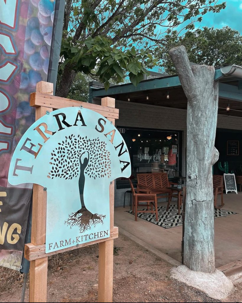 Outdoor sign for Terrasana Farm+Kitchen with a tree and roots, mounted on wood, cafe seating visible in background, Vintage Cork, Ben Wheeler, Texas