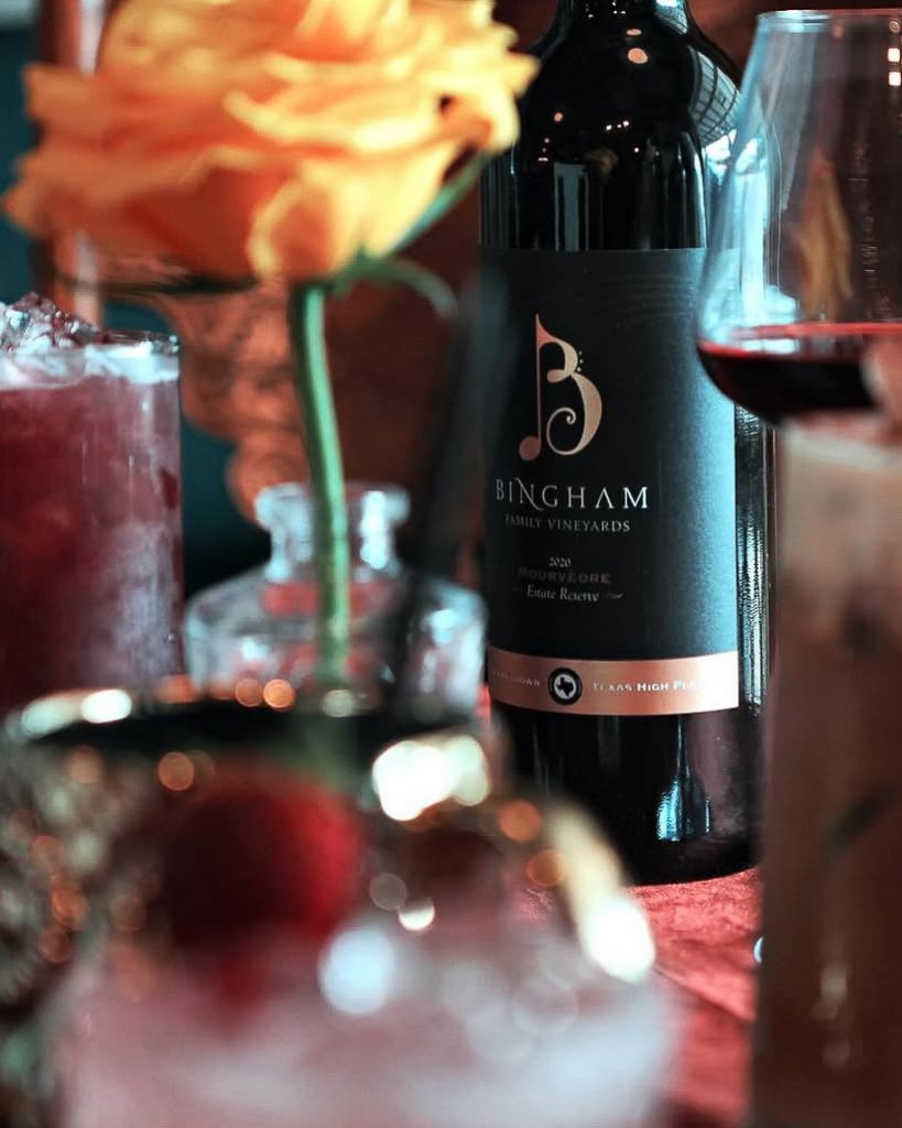 Bottle of Bingham Family Vineyards Mourvèdre on a table, with a pink flower in the foreground and a glass of red wine nearby, Vintage Cork, Ben Wheeler, Texas