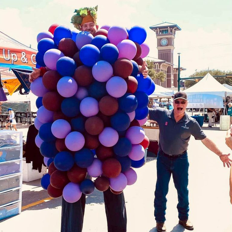 Cliff Bingham beside a giant grape character at GrapeFest in Historic Downtown Grapevine, with the Grapevine clock tower in the background.