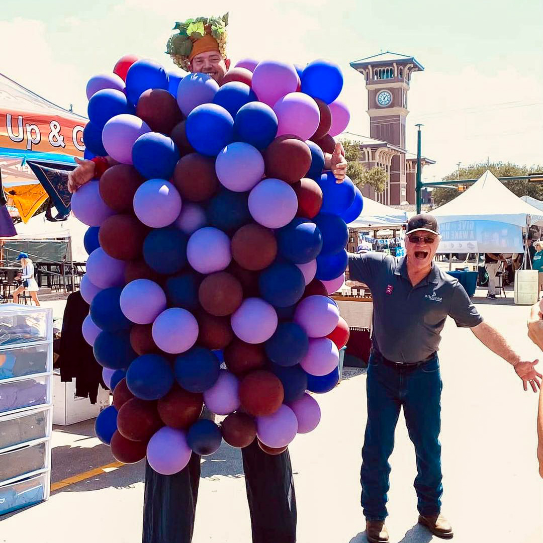 Cliff Bingham beside a giant grape character at GrapeFest in Historic Downtown Grapevine, with the Grapevine clock tower in the background.