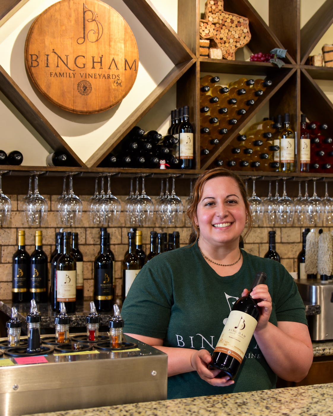 Emmalee at Bingham Family Vineyards Grapevine tasting room behind the bar and holding a bottle of Bingham wine.